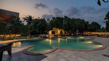 A swimming pool with a curved edge, surrounded by a tiled deck, and a small building with a light fixture in the background.