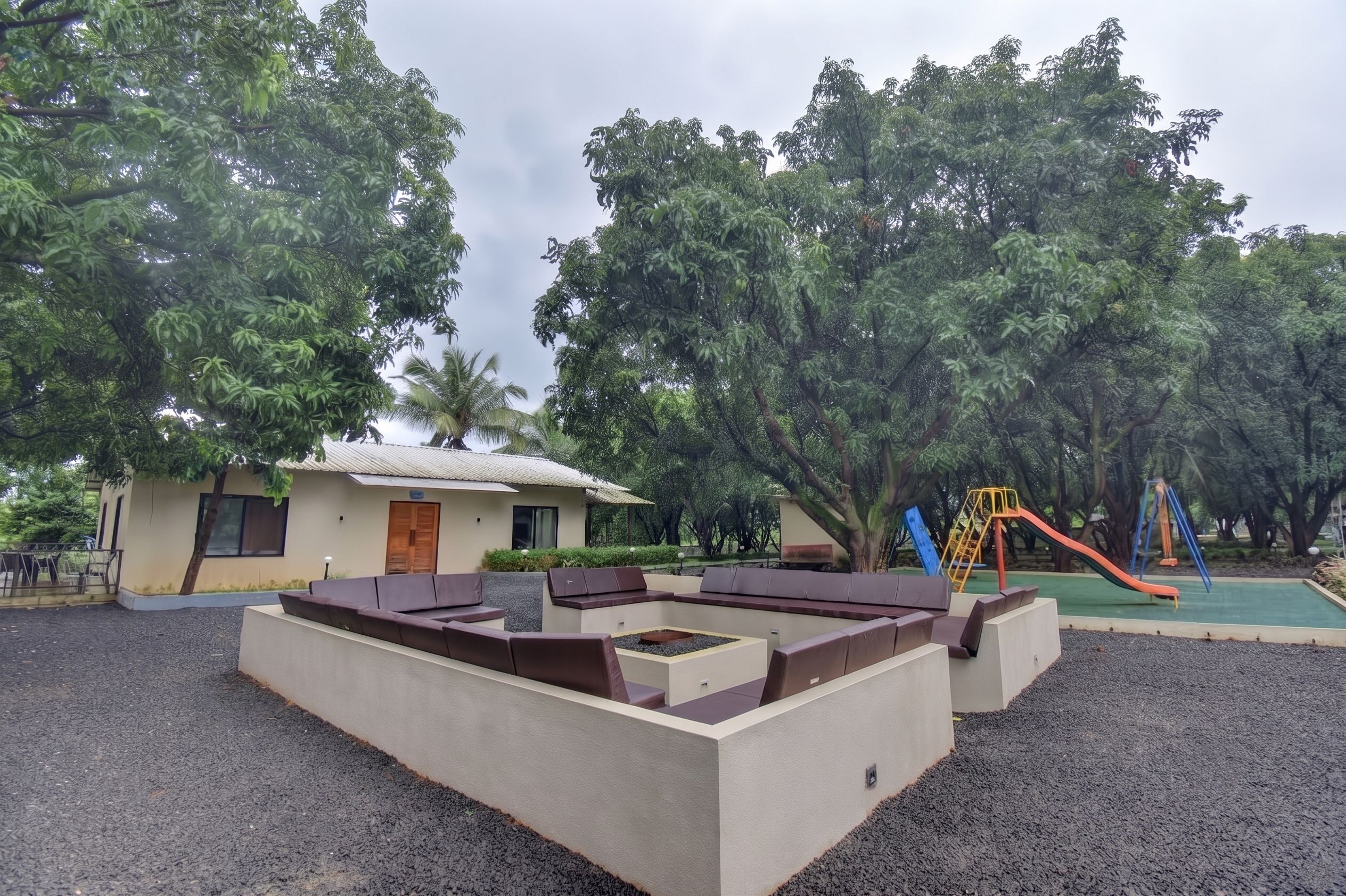A playground with a slide and swings, surrounded by trees and a building with a sloped roof.