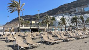 A beach with rows of sun loungers and palm trees, a building with multiple floors in the background, and a mountainous landscape. - Sunny Seaview Modern apt - Pool and Beach Access (Mogan)