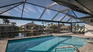 A clear swimming pool with a glass enclosure, surrounded by palm trees and a view of a residential area. - Relaxing Retreat, Heated Pool,  Gulf Access NEW (Cape Coral)