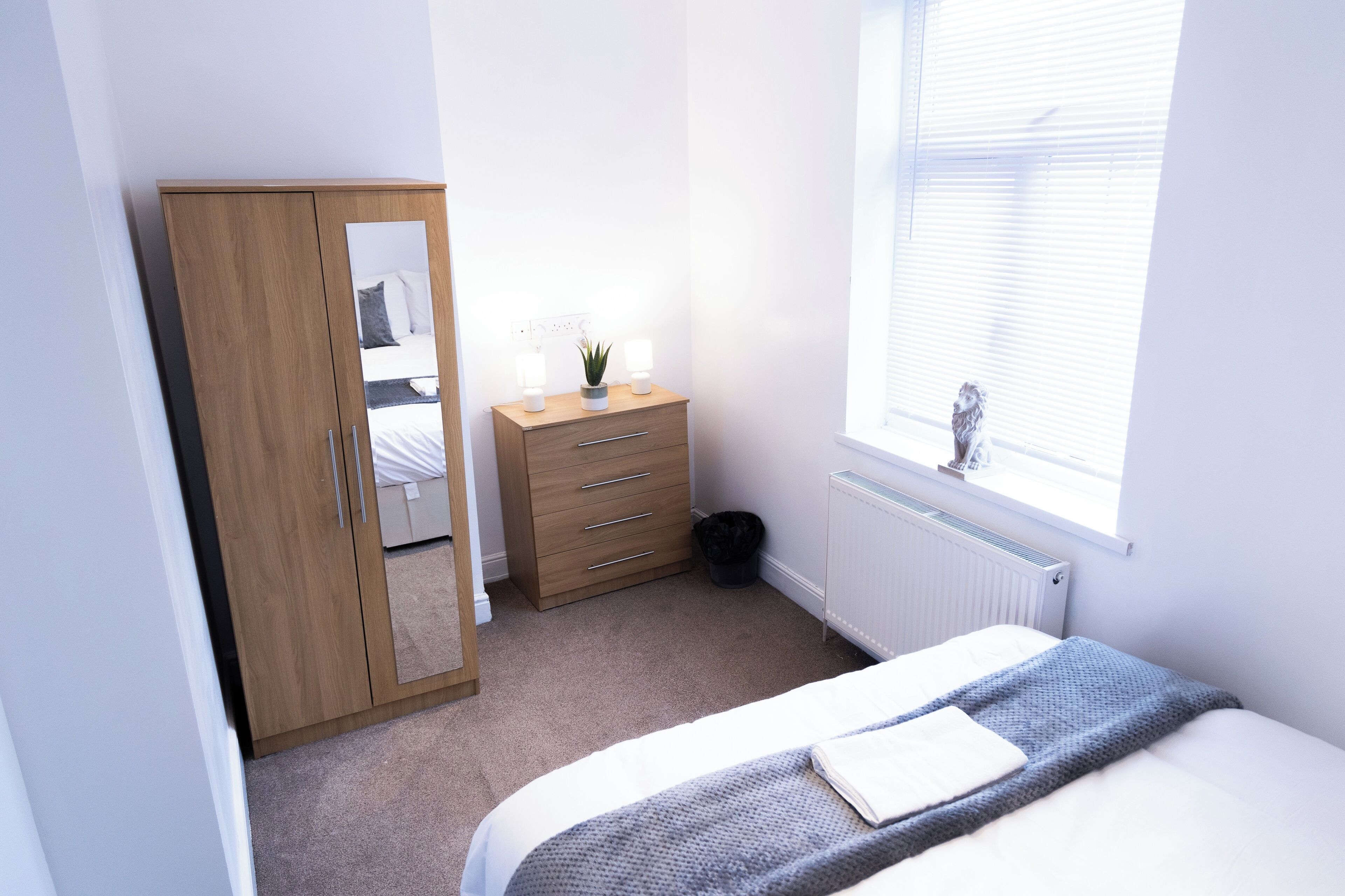 Un dormitorio con cama, armario de madera, mesita de noche con lámpara y planta, radiador y ventana con persianas.