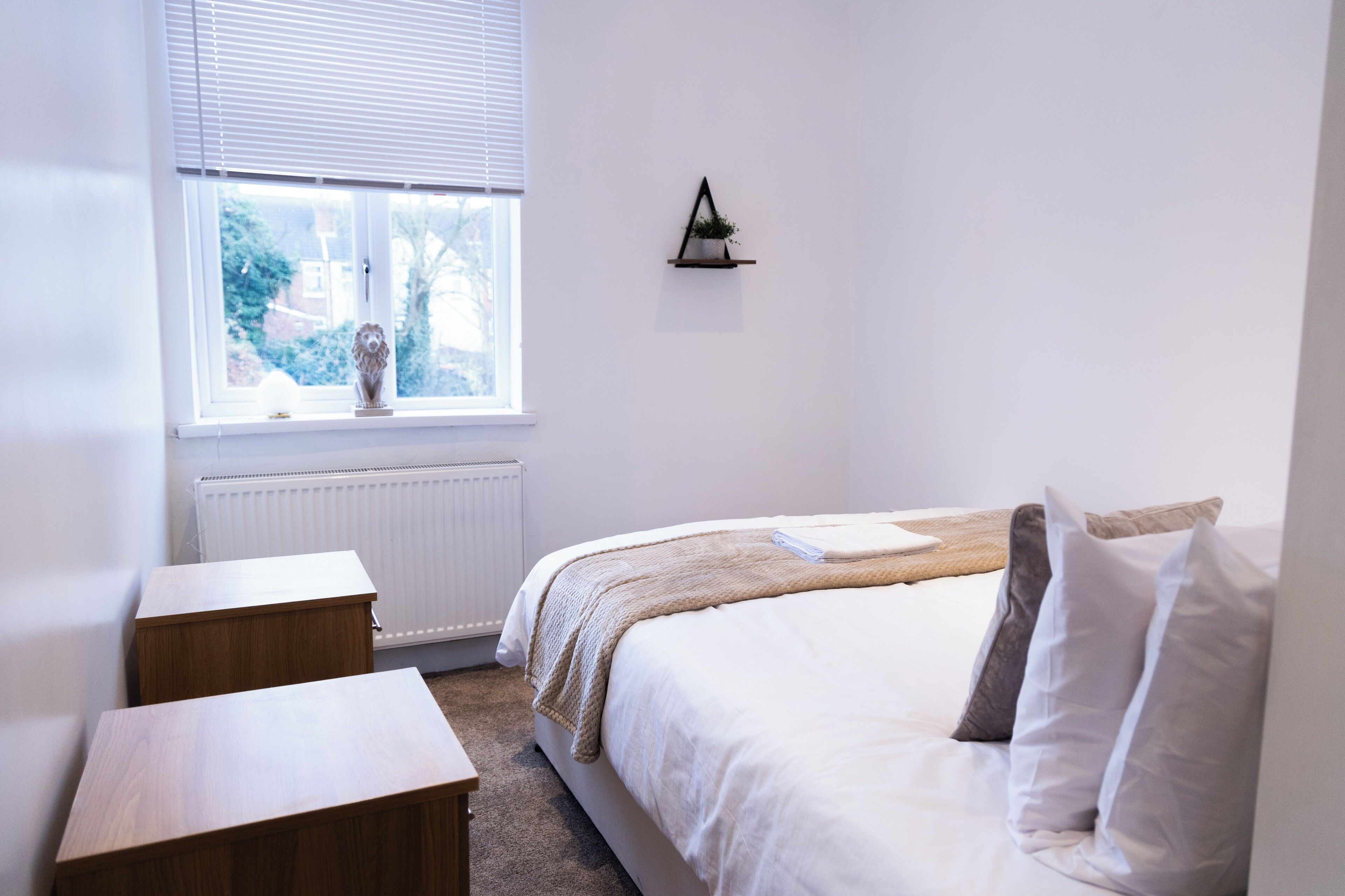 Un dormitorio con cama, mesitas de noche, ventana con vistas, radiador y estante en la pared con una planta.