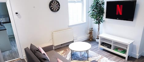 A modern living room with a sofa, a coffee table, a dartboard, a radiator, a plant, a TV, and a bookshelf.