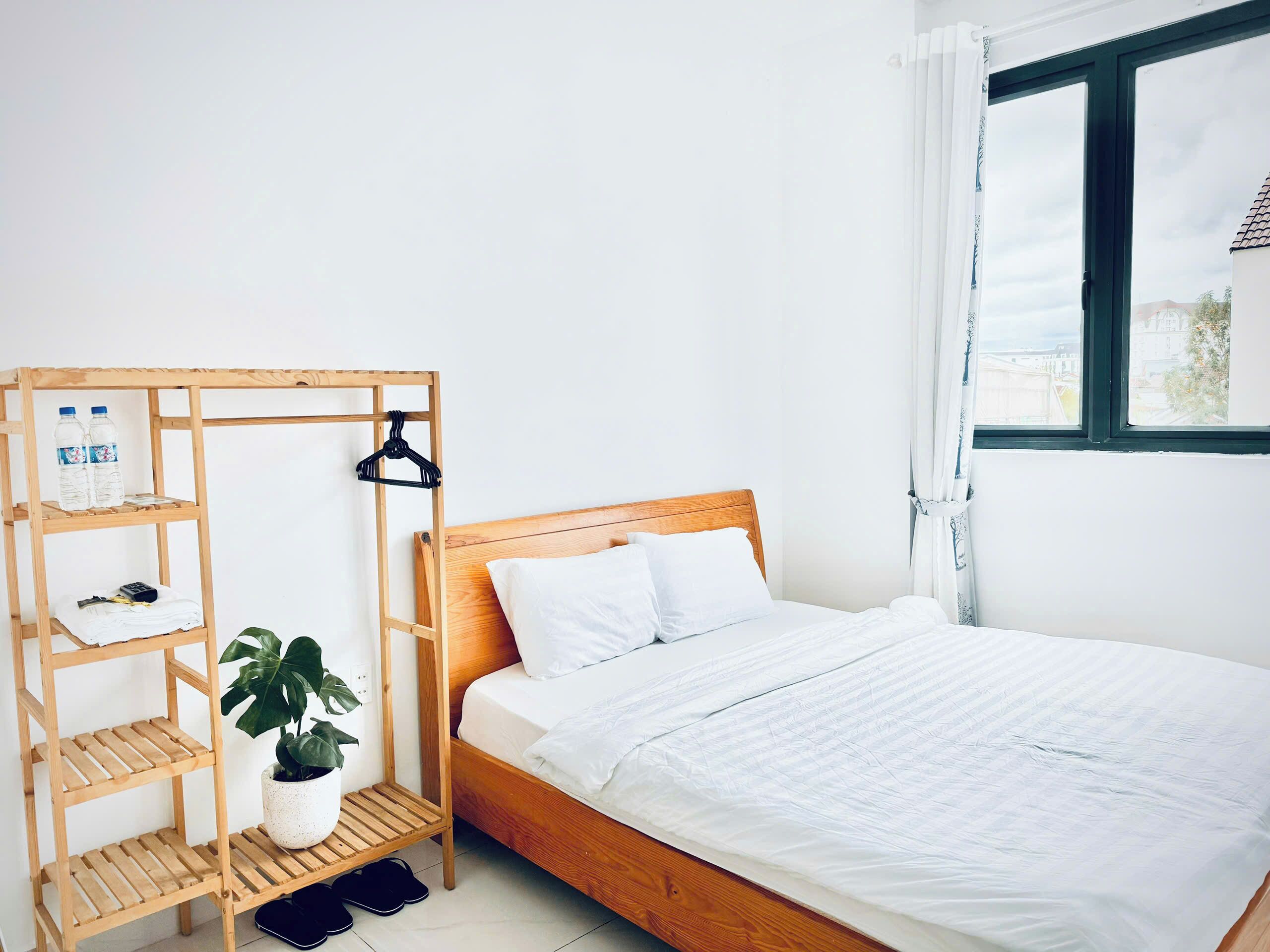 A neatly made bed with white bedding, a wooden headboard, and a wooden shelf unit with a plant and bottles beside it.