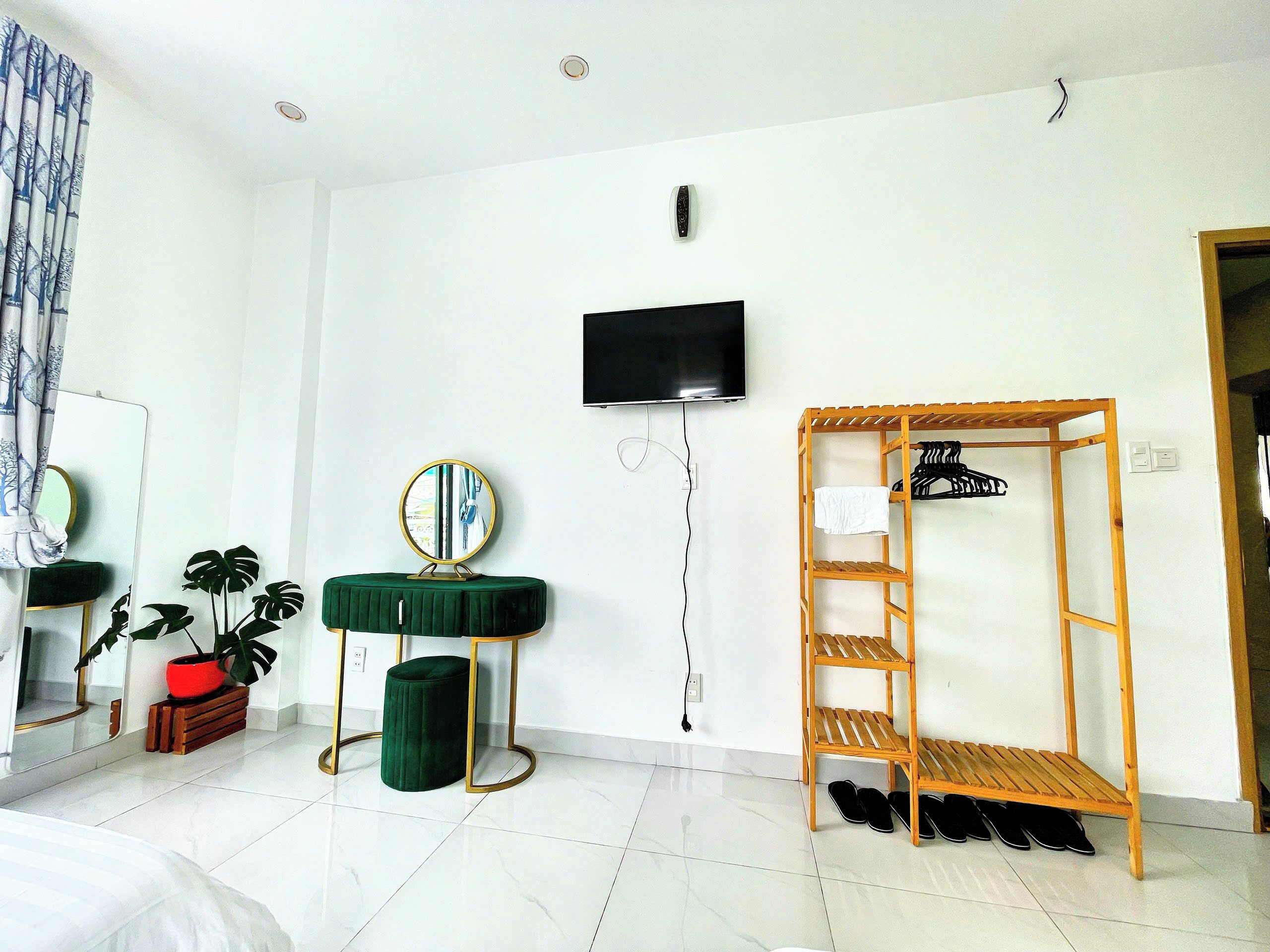 A hotel room with a bed, a TV mounted on the wall, a wooden shoe rack, a green ottoman, and a mirror with a plant.