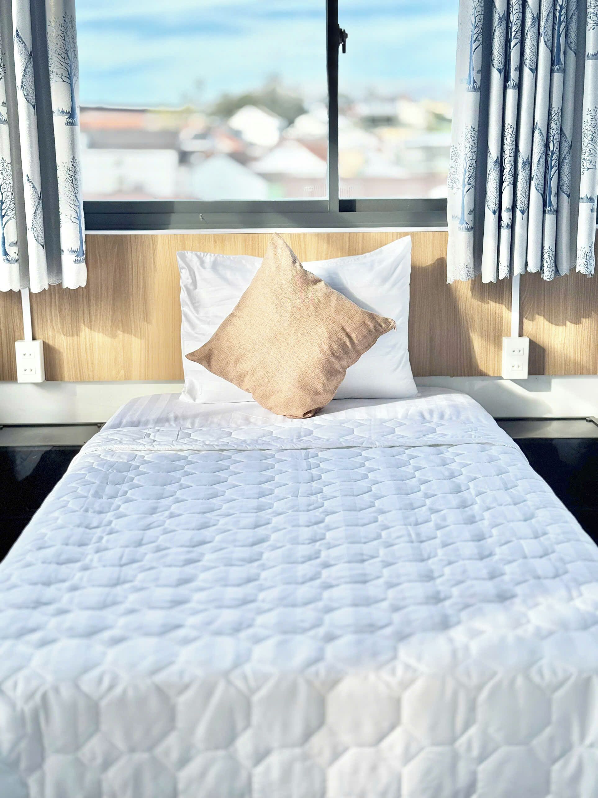 Una cama bien hecha con colcha blanca y almohada color beige, ubicada frente a una ventana con cortinas.