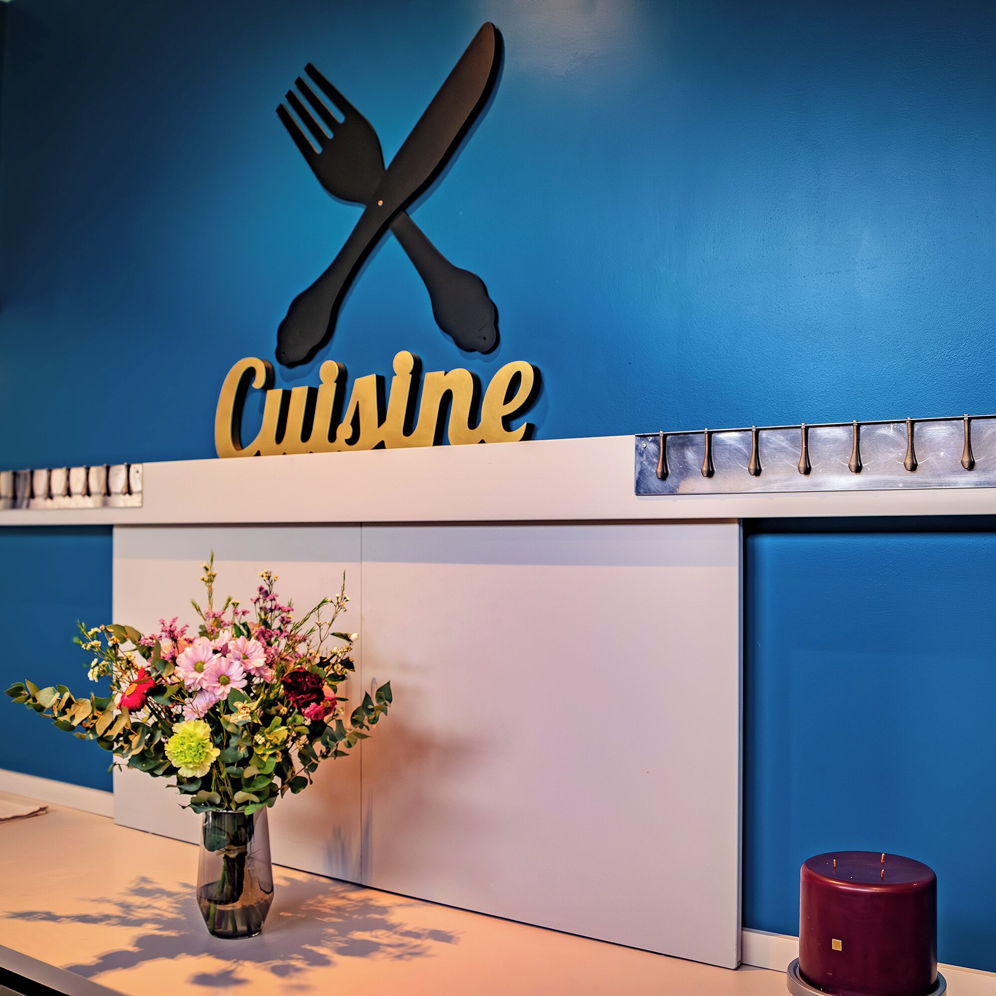 Un comptoir d’accueil avec un mur bleu, une enseigne indiquant « Cuisine » au-dessus, un vase avec des fleurs et un chandelier.