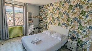 A bedroom with a bed, a nightstand, a radiator, a shelf, and a window with a view of buildings. - Hôtel de l'Abbaye  (Cluny)