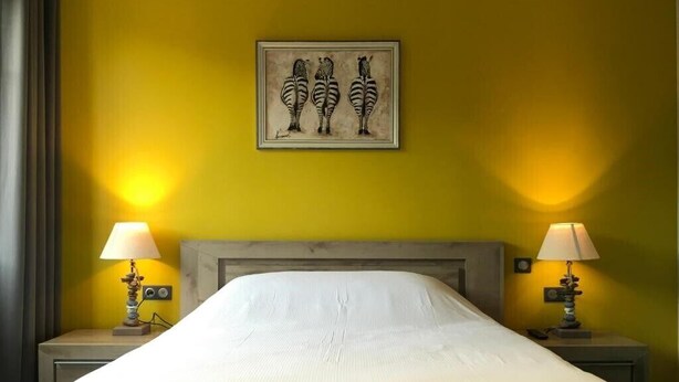 A hotel room with a bed, two bedside lamps, a wall-mounted painting of zebras, and a yellow wall. - Hôtel de l'Abbaye  (Cluny)