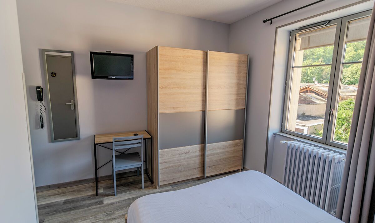 A bedroom with a bed, a desk, a wardrobe, a TV, and a window with a view.