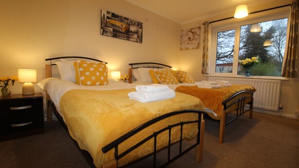 A double room with two beds, each with a yellow polka-dotted bedspread and matching pillows. There is a window with curtains, a radiator, and a wall-mounted picture. - Keswick House Chesterfield (Chesterfield)