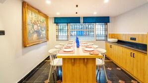A dining area with a long wooden table set for multiple people, white chairs, a large painting on the wall, a window with a blue valance, and a kitchen area with wooden cabinets and a sink.