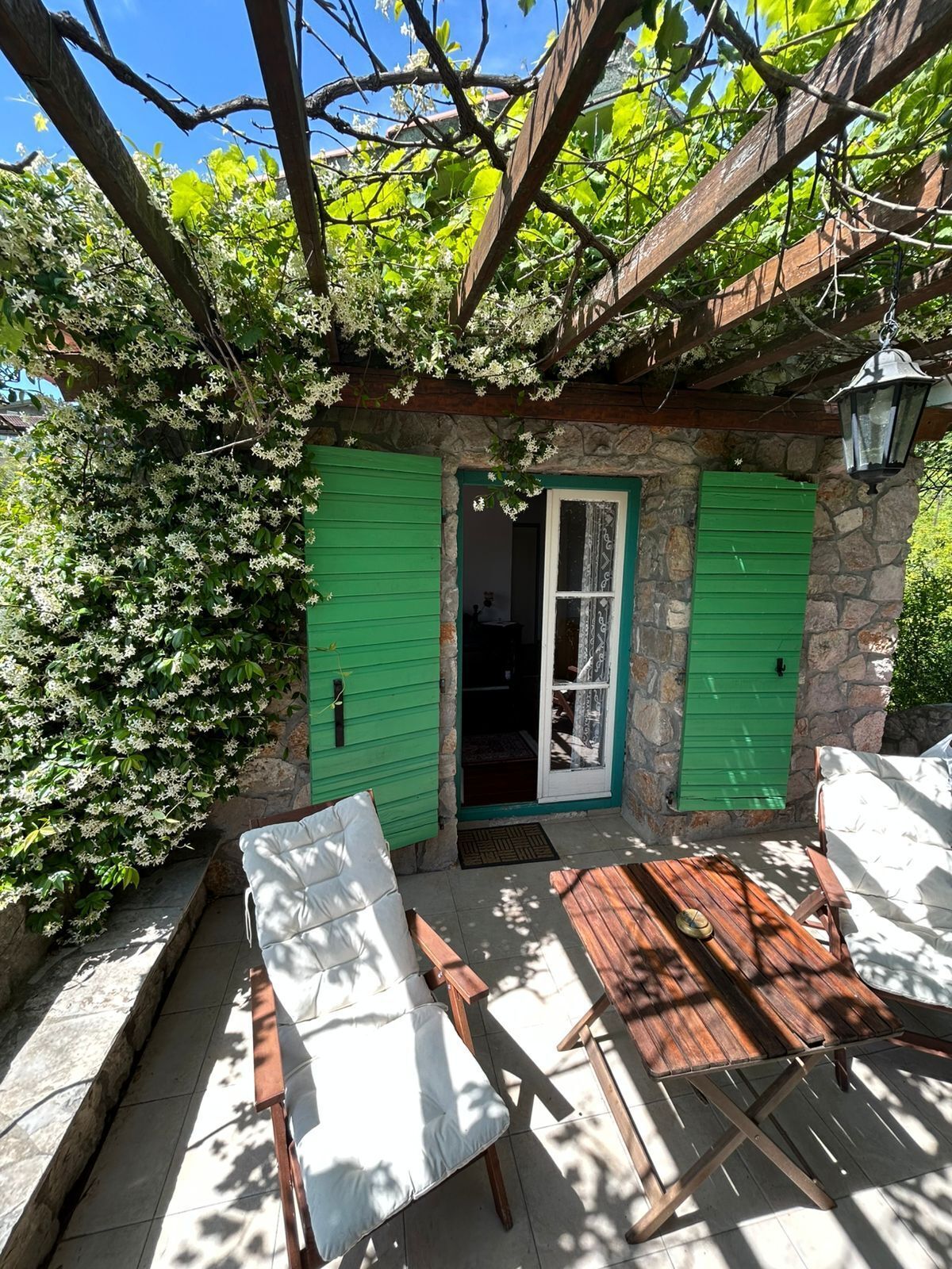 A stone cottage with green shutters, a wooden table, and a white cushioned bench.