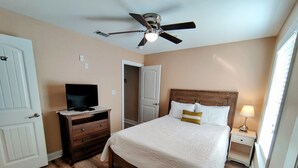 Un dormitorio con una cama, un televisor sobre una cómoda, un ventilador de techo y una mesita de noche con una lámpara.