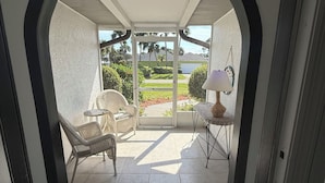 A covered patio area with a view of a residential street, a wicker chair, a small table with a lamp, and a welcome mat. - Coastal Getaway with game room pool hot tub & inlaw suite in heart of Sebastian (Sebastian)