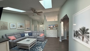 A spacious living room with a grey sectional sofa, a coffee table, and a ceiling fan. There are framed pictures on the walls and a large window providing natural light. - Coastal Getaway with game room pool hot tub & inlaw suite in heart of Sebastian (Sebastian)