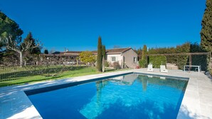 A rectangular swimming pool with clear blue water, surrounded by a paved deck. There are lounge chairs and a small table on the deck. In the background, there are houses and a fence with trees. - Beautiful home in Graveson (Graveson)