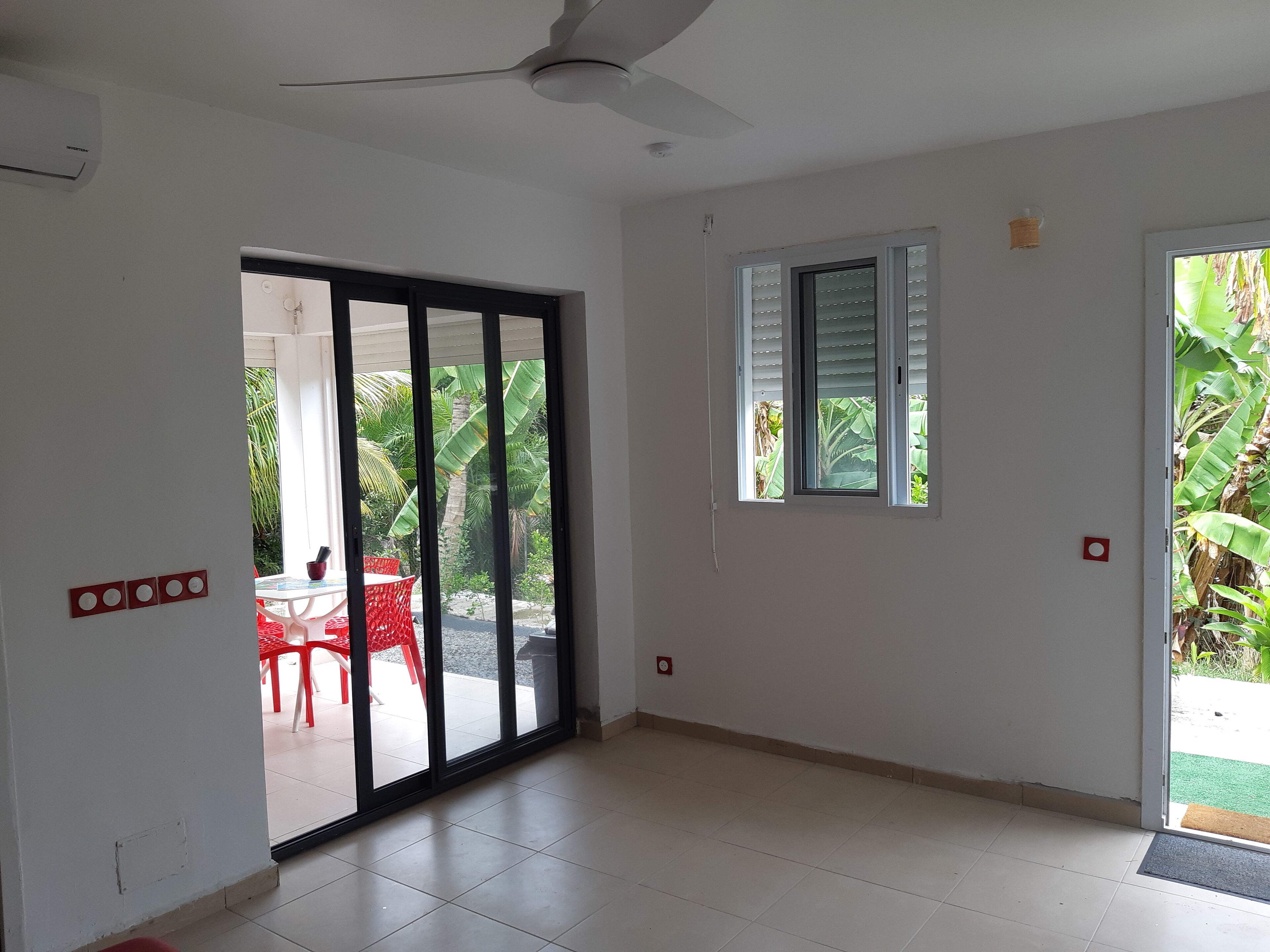 A room with a sliding glass door leading to an outdoor area with a red chair and table, a ceiling fan, a window with blinds, and a small red light switch on the wall.