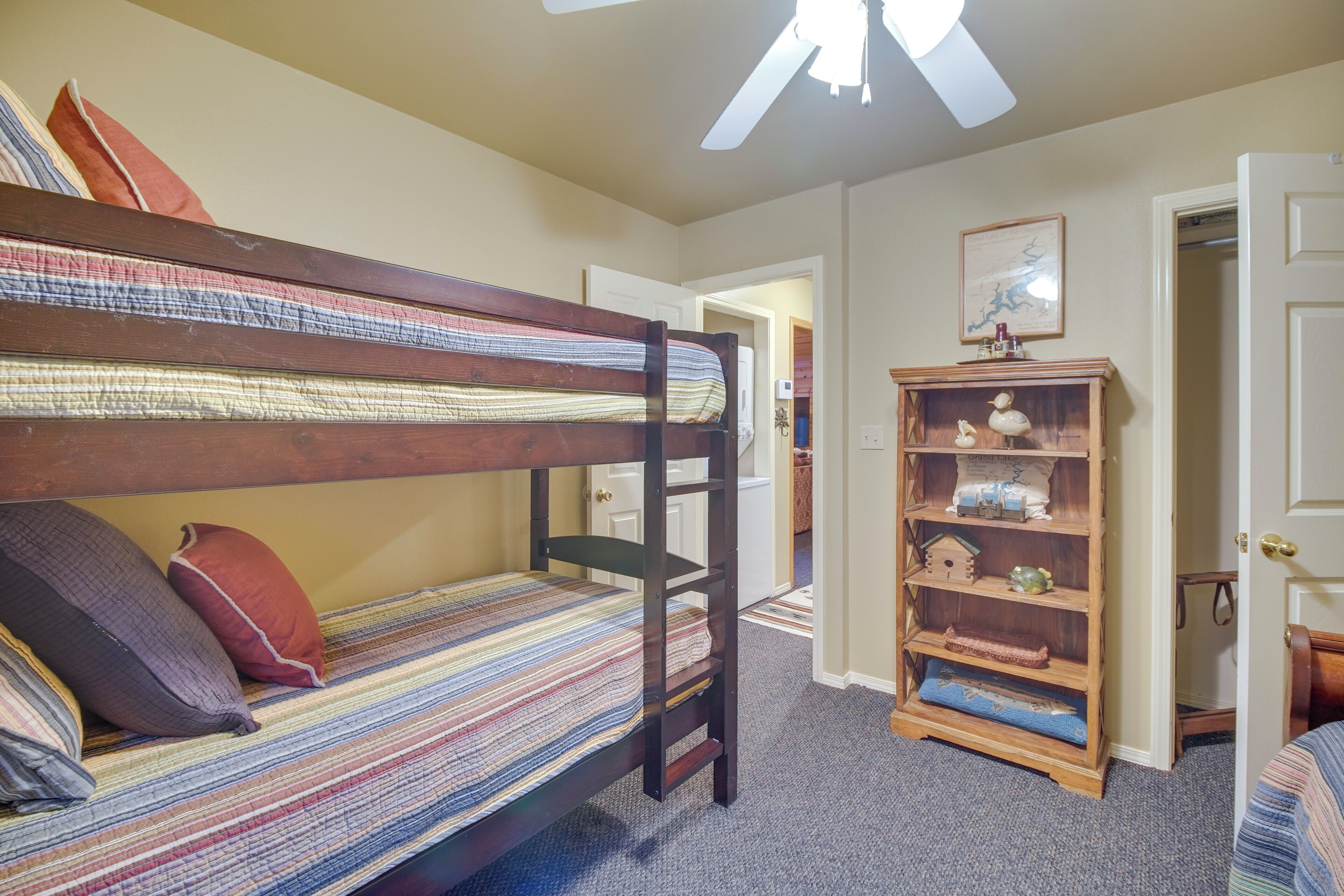 A bunk bed with striped bedding, a wooden bookshelf, and a ceiling fan in a room with beige walls and carpet.