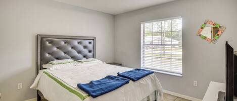 A bedroom with a tufted headboard, a bed with white bedding and blue towels, a window with blinds, a mirror, and a TV on a stand.