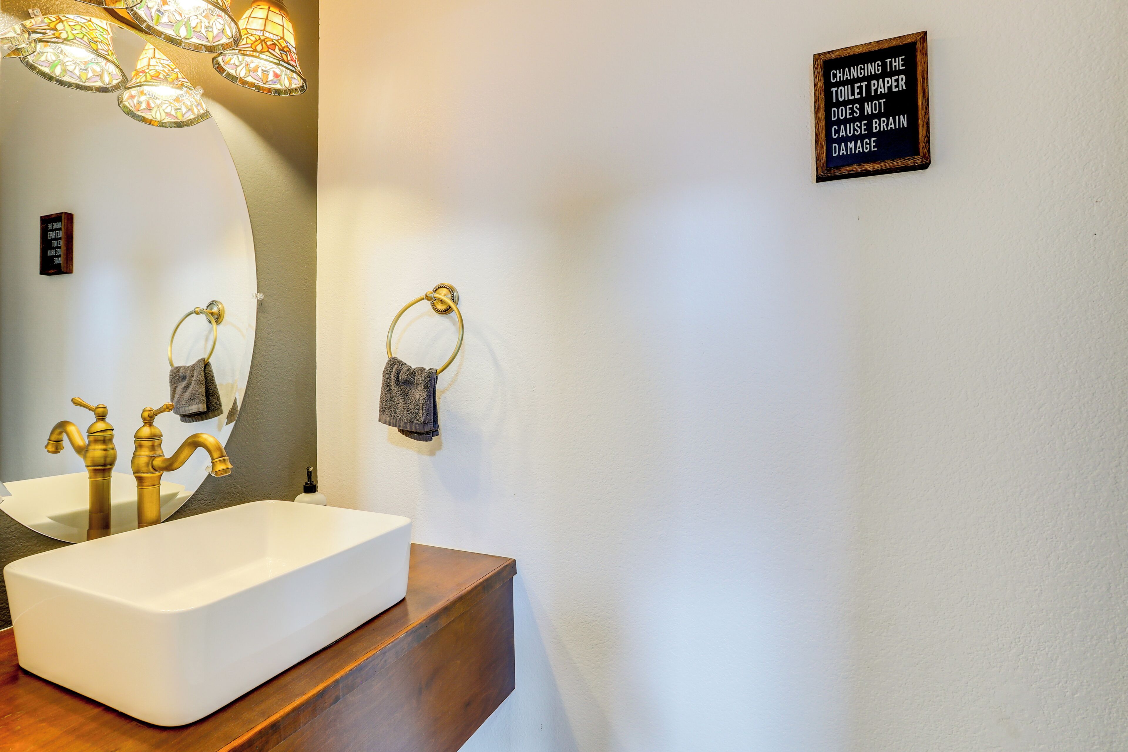 Ein Badezimmer mit einem weißen Waschbecken, goldenen Wasserhähnen und einem Spiegel. An der Wand befindet sich ein Handtuchhalter und ein Schild mit der Aufschrift "Toilettenpapier wechseln schadet nicht dem Gehirn".