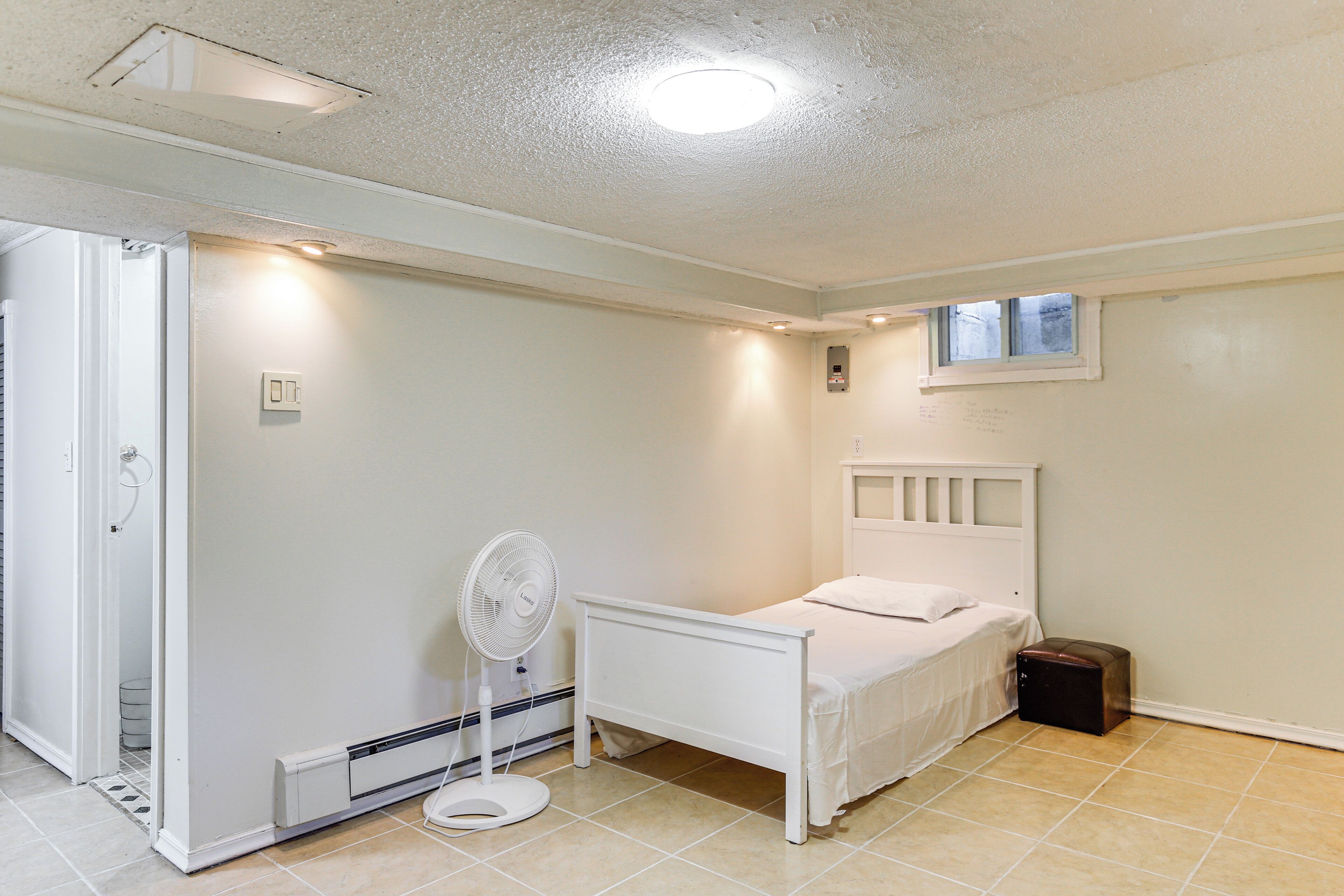 Une chambre avec un lit, un ventilateur sur pied, un radiateur et une petite fenêtre.