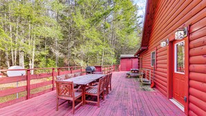 Una terraza de madera con una mesa y sillas, rodeada por una cerca roja y un cobertizo pequeño en un área boscosa.