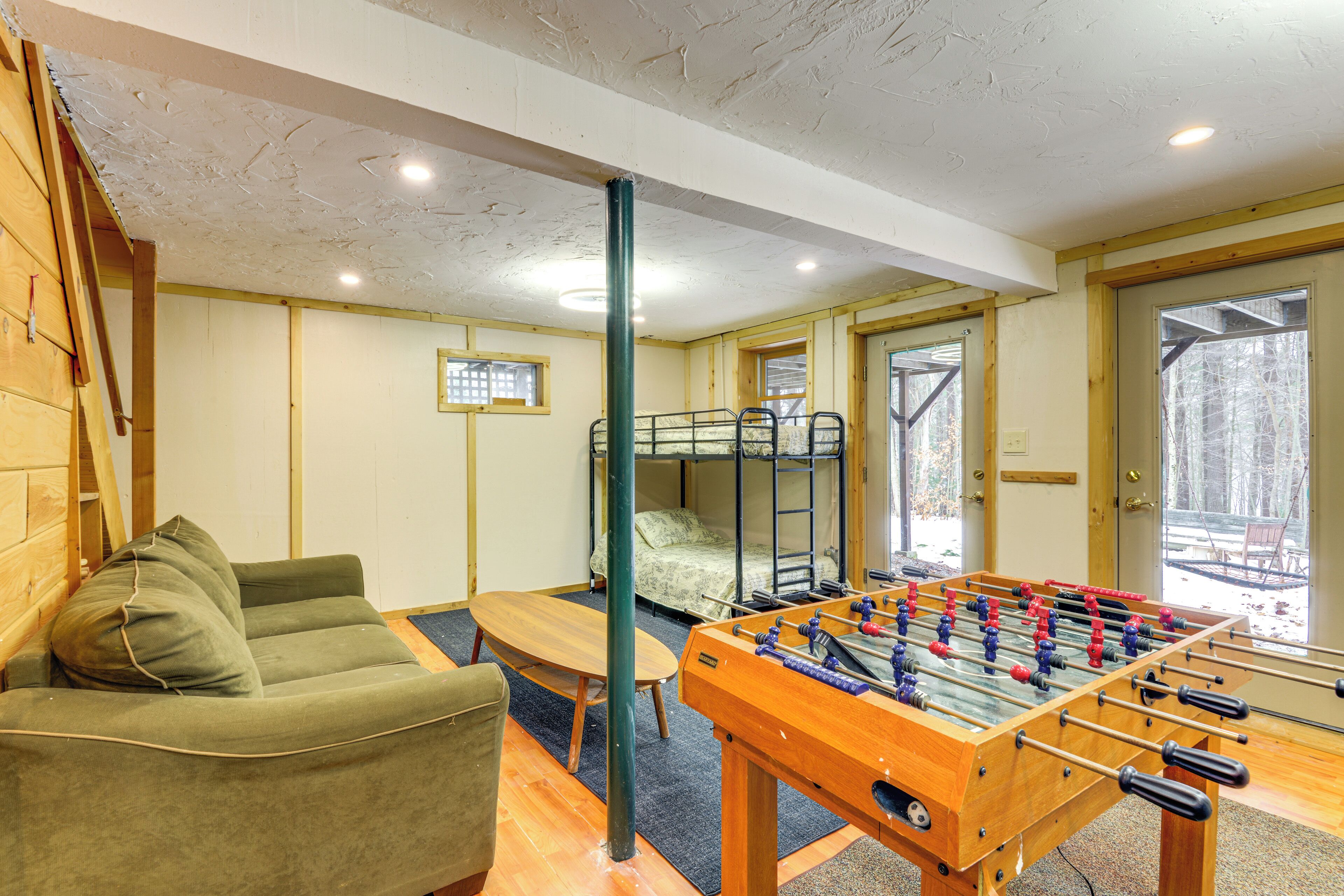 A room with a foosball table, bunk beds, a sofa, and a window with a view of trees.