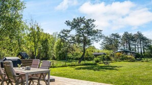 A wooden deck with outdoor furniture, a grill, and a grassy lawn with trees and a small building in the background.