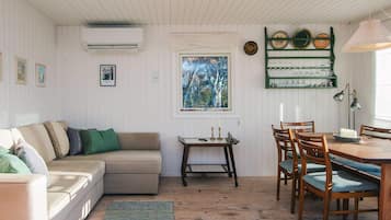 A cozy living room with a sofa, a dining table with chairs, a window with a view of trees, and a wall-mounted shelf with decorative plates.
