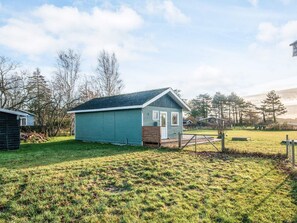 A small, single-story building with a green exterior and a sloped roof, surrounded by a grassy yard and trees. - 4 Person Holiday Home in Knebel (Knebel)