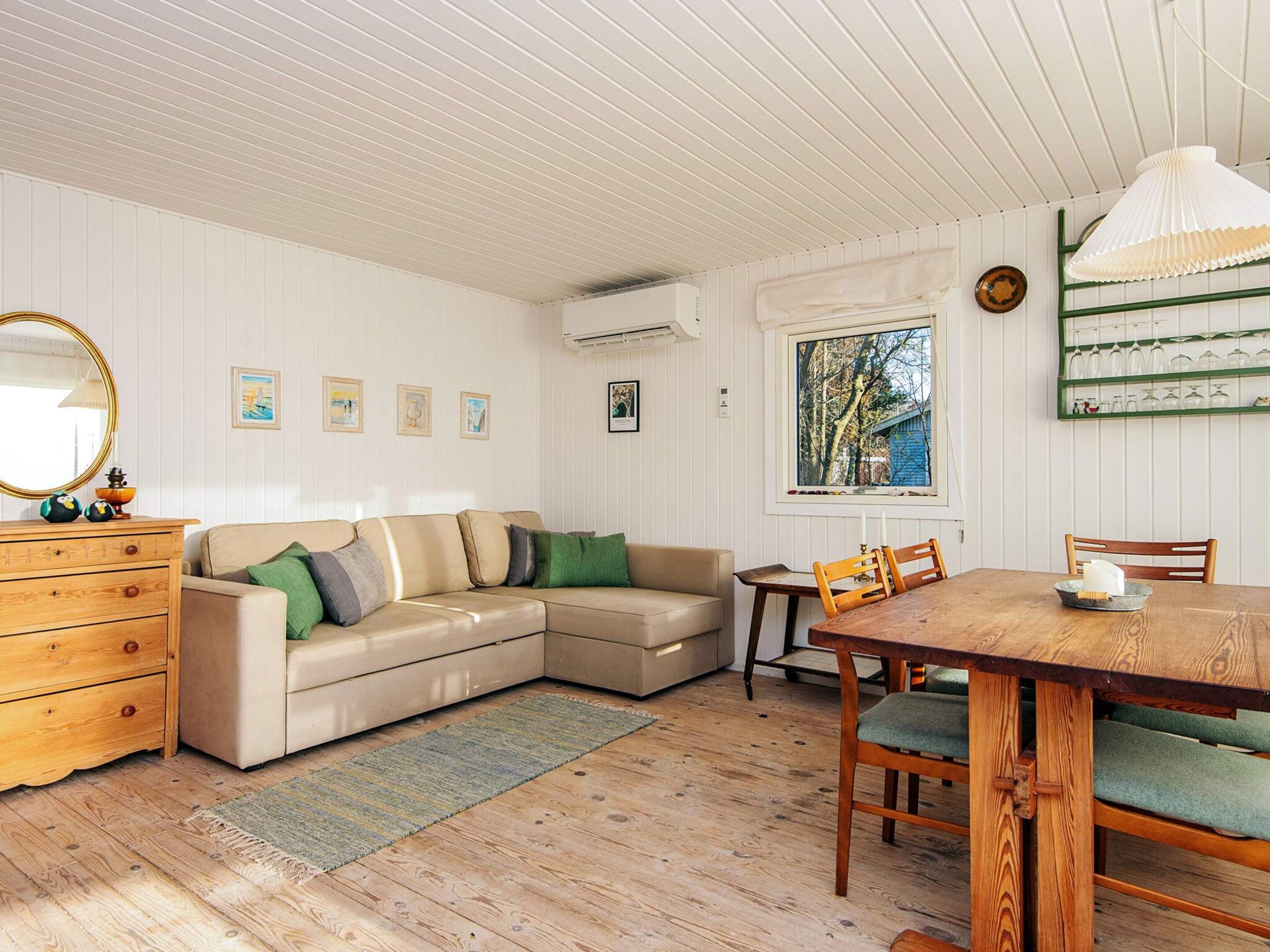 A cozy room with a wooden floor, a beige sectional sofa with green cushions, a wooden dresser, a dining table with chairs, and a window with a view of trees.