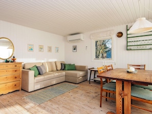 A cozy room with a wooden floor, a beige sectional sofa with green cushions, a wooden dresser, a dining table with chairs, and a window with a view of trees. - 4 Person Holiday Home in Knebel (Knebel)