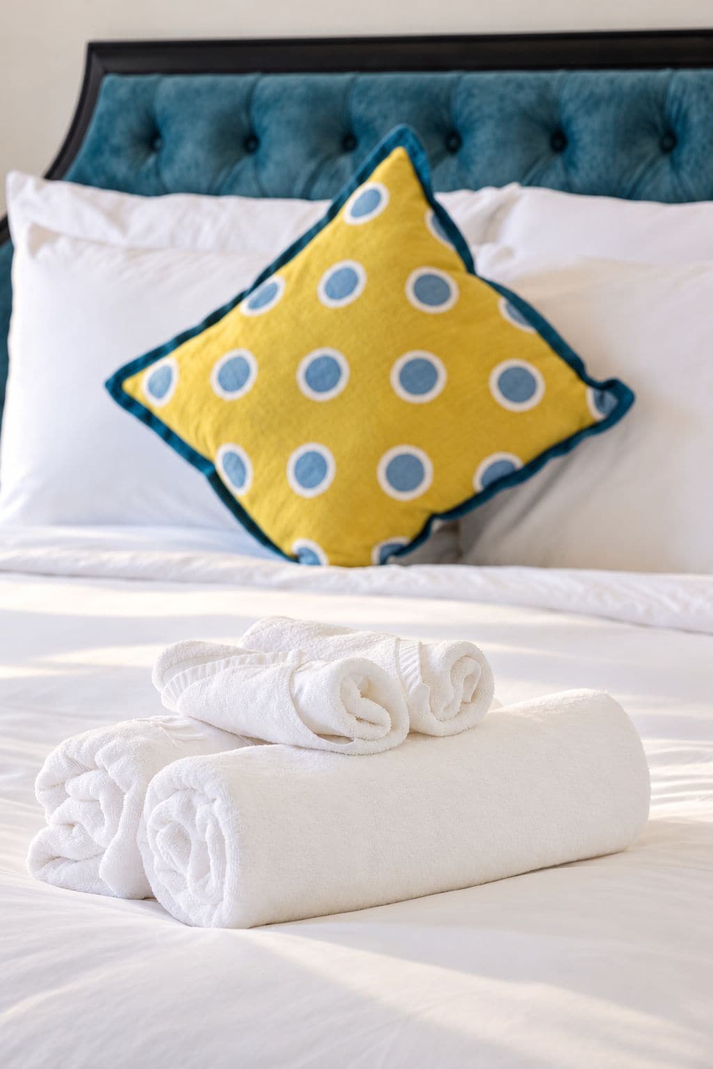 A neatly made bed with white linens and a rolled white towel, a blue tufted headboard, and a yellow pillow with blue polka dots.