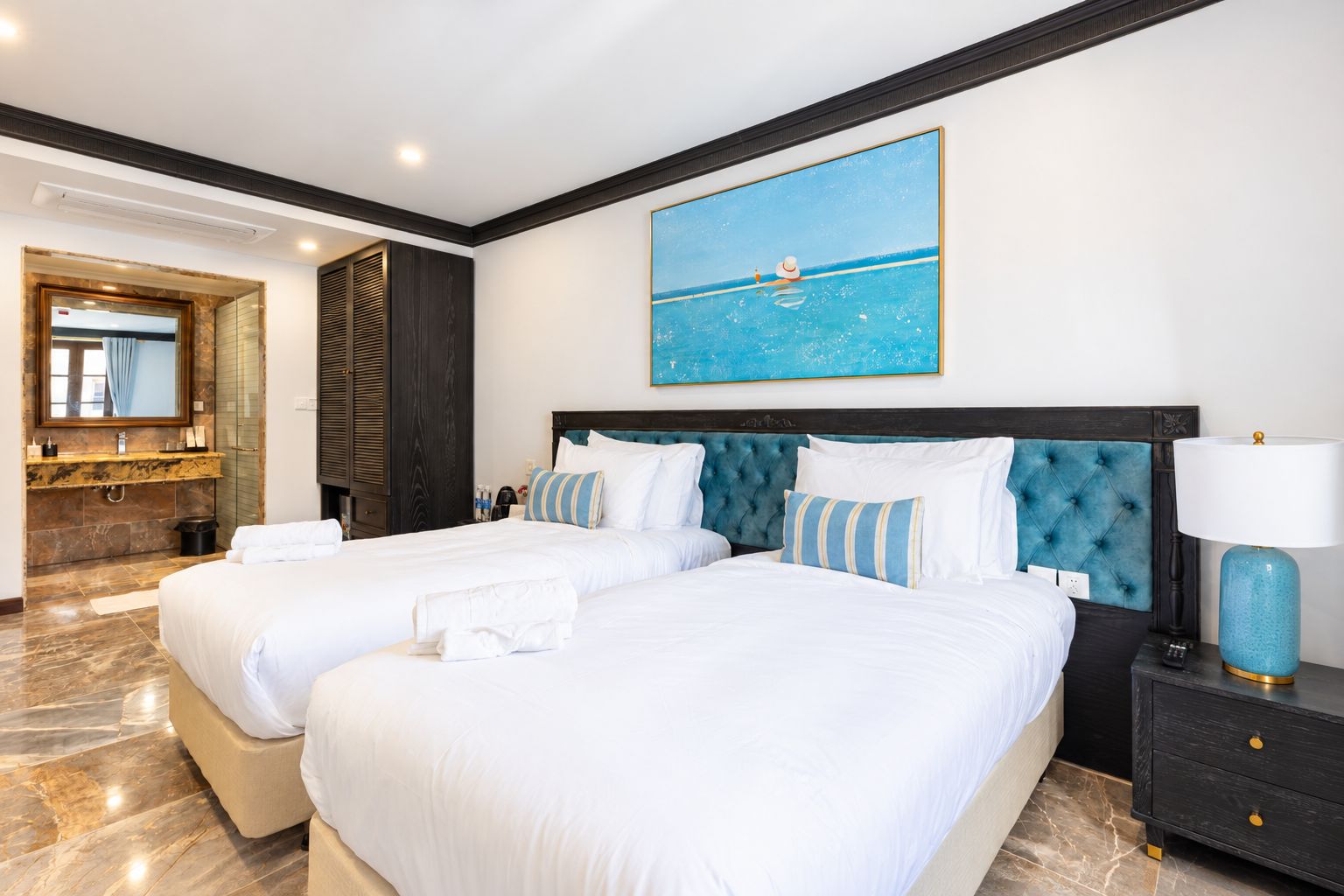 A hotel room with two beds, a large painting of a beach scene, a dark headboard, and a nightstand with a lamp.