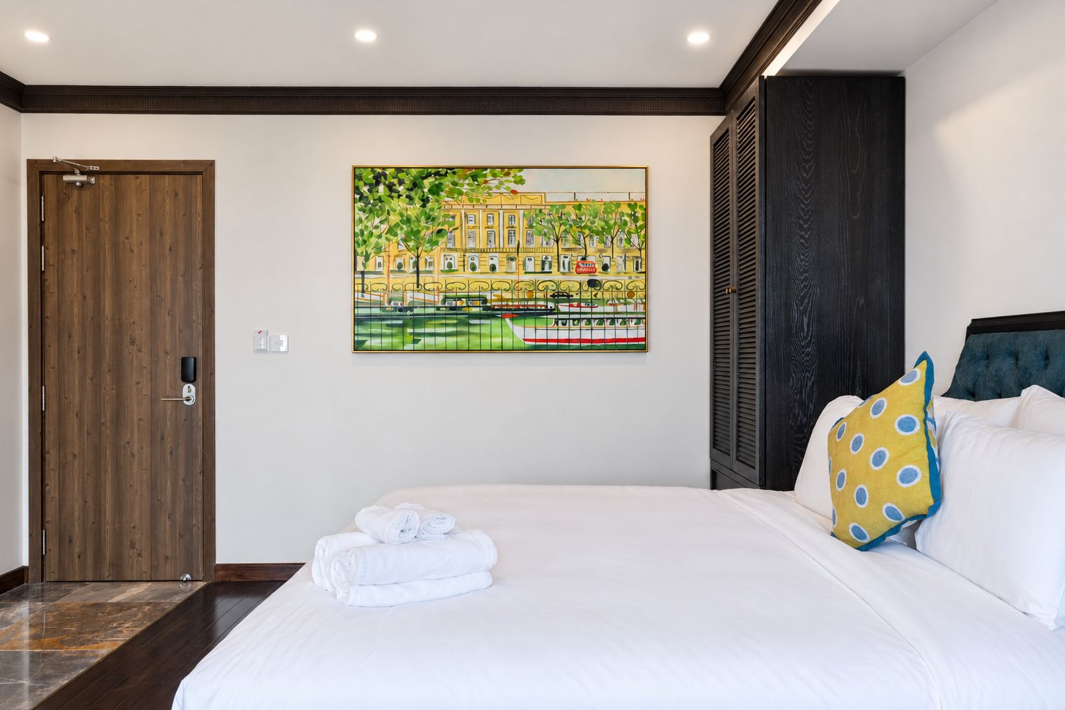 A hotel room with a large bed, a painting of a cityscape, a wooden door, and a dark-colored headboard.