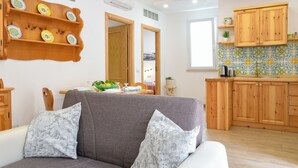 Una sala de estar con un sofá gris, dos almohadones estampados, un estante de madera con platos decorativos, y una cocina con armarios de madera y salpicadero de azulejos.