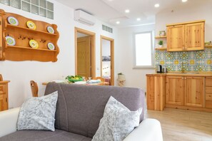 Un salon avec un canapé gris, deux coussins à motifs, une étagère en bois ornée d’assiettes décoratives, et une cuisine avec des meubles en bois et un dosseret carrelé.