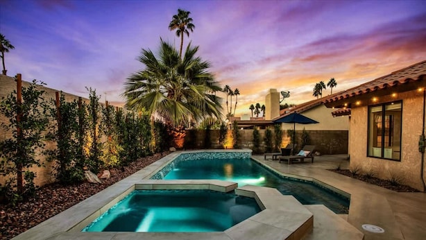 A swimming pool with a stone border, surrounded by palm trees and a sunset sky. - Paradise at Waring Place (Palm Desert)