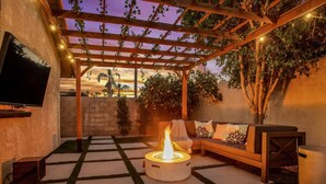 A patio area with a fire pit, outdoor seating, a TV mounted on the wall, and a pergola with string lights. - Paradise at Waring Place (Palm Desert)
