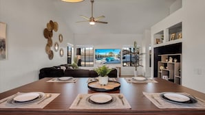 A dining area with a long table set for four, a ceiling fan, and a large window with a view of a pool. - Paradise at Waring Place (Palm Desert)