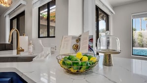 A modern kitchen with a marble countertop, a gold faucet, a bowl of limes, and a view of a swimming pool. - Paradise at Waring Place (Palm Desert)