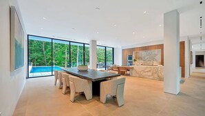 Ein geräumiger Essbereich mit großem Tisch und Stühlen, eine Küche mit Marmorarbeitsplatten und große Fenster mit Blick auf einen Swimmingpool und Grünflächen.