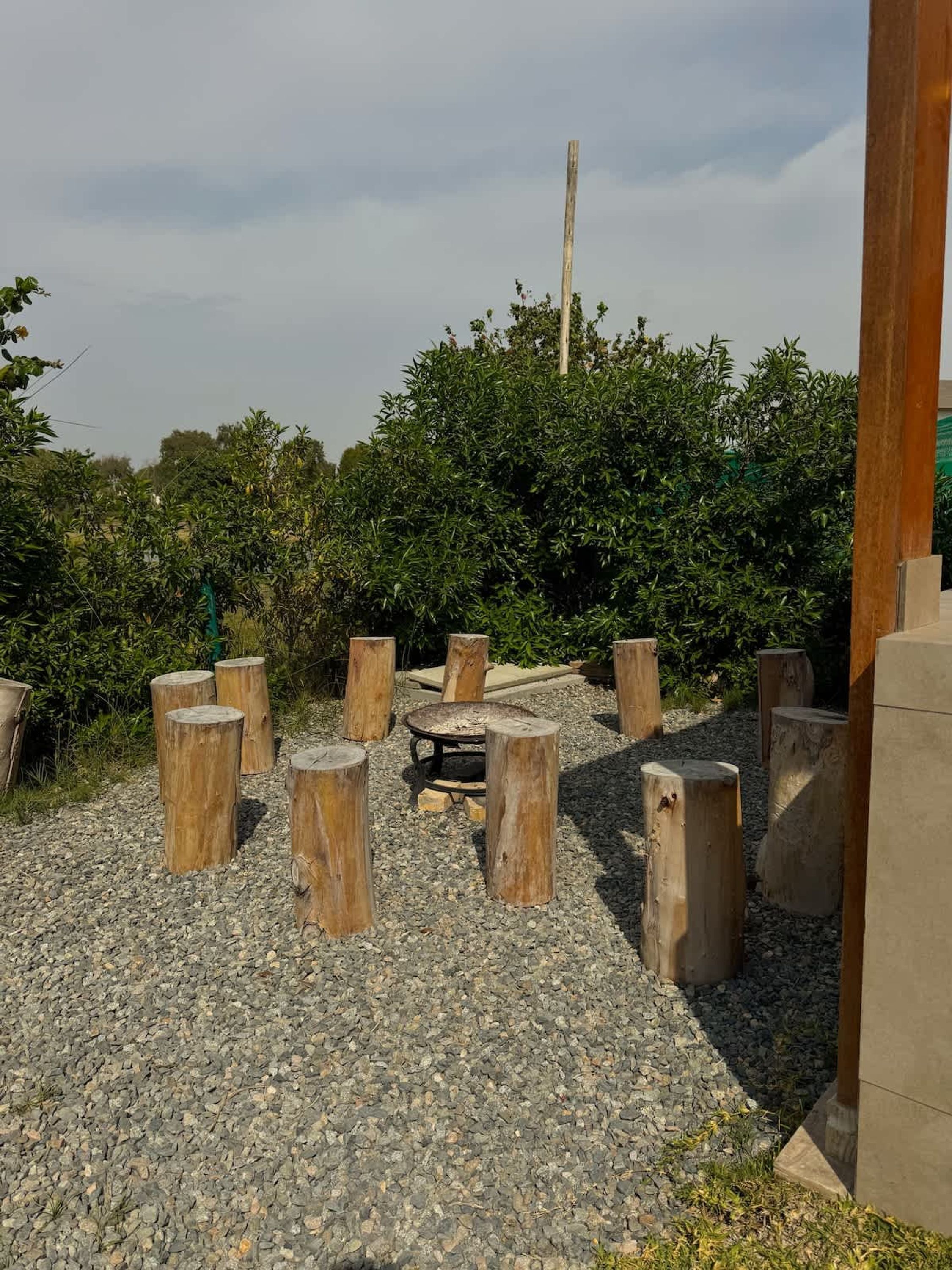 A gravel area with wooden posts arranged in a circle, surrounded by greenery and a tall wooden pole.