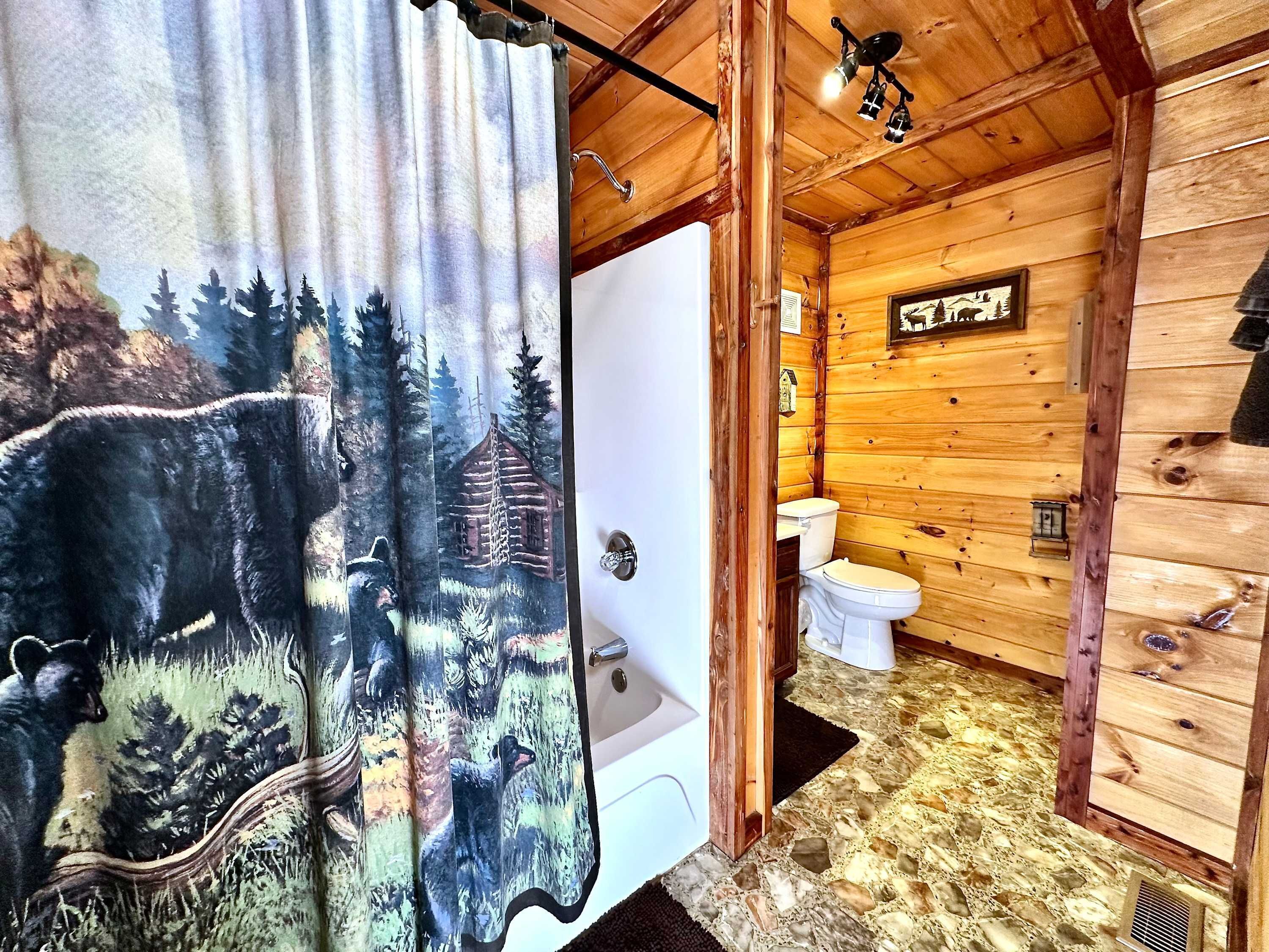 A rustic wooden cabin bathroom with a wooden floor, a white toilet, and a shower curtain depicting bears and trees.