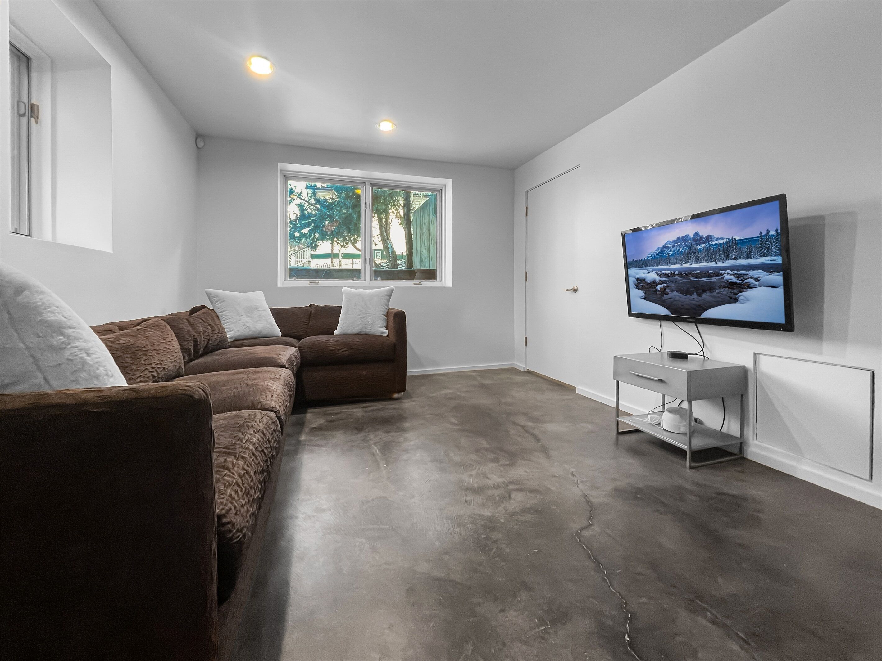 A modern living room with a sectional sofa, a flat-screen TV, and a window with a view of trees.
