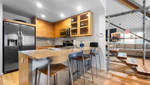 Eine moderne Küche mit Edelstahlkühlschrank, Mikrowelle und Holzschränken. Drei Barhocker und eine Arbeitsplatte aus Granit ergänzen das Bild.