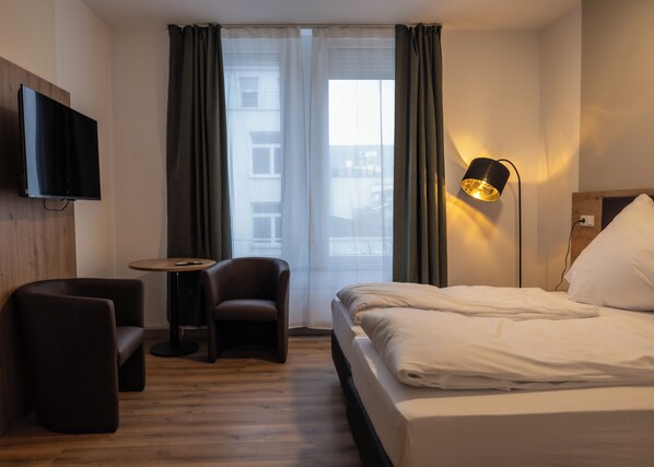 Habitación de hotel con cama, una silla, una mesita, televisor montado en la pared y ventana con cortinas.