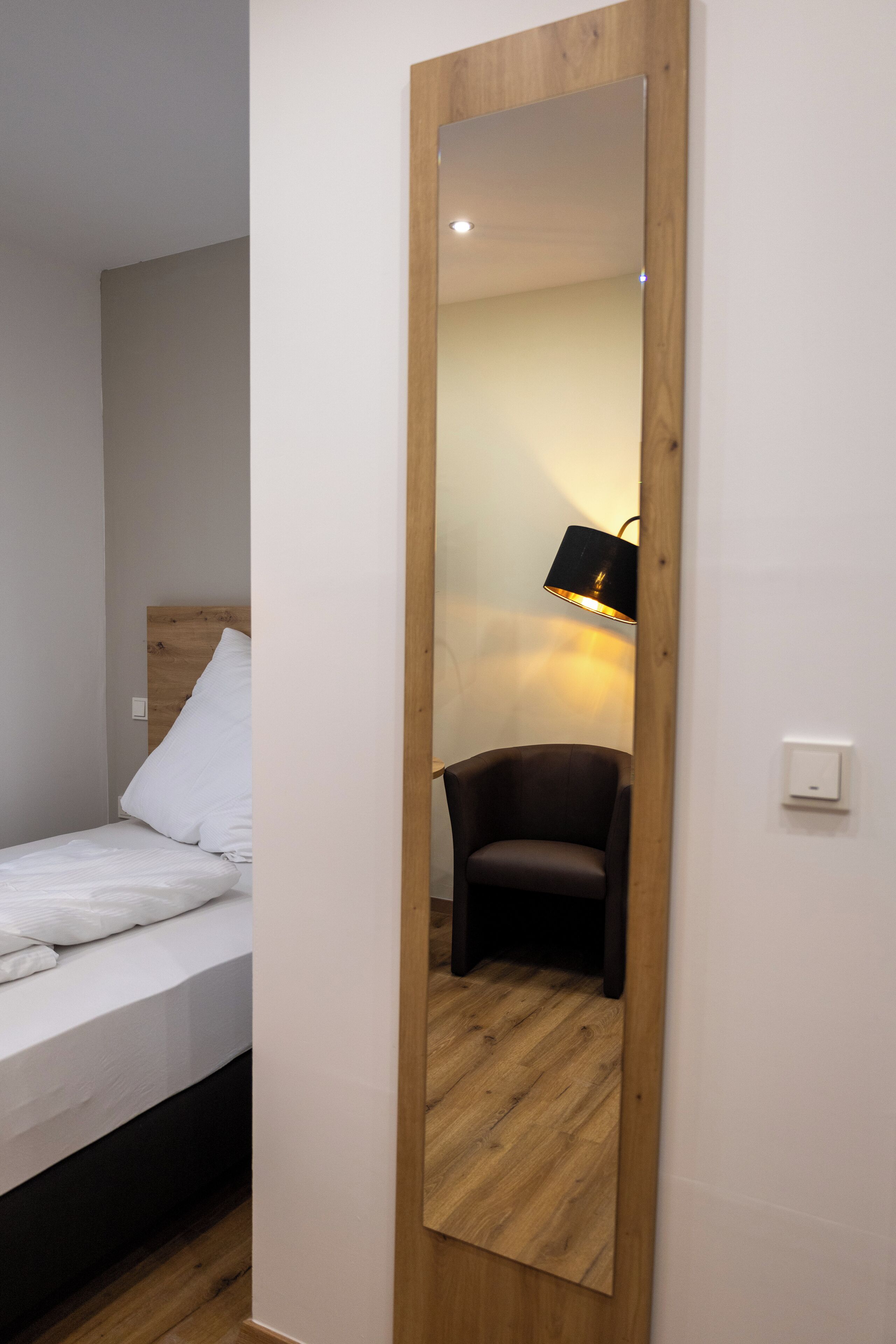 A bedroom with a wooden headboard bed, a large mirror, a chair, and a bedside lamp.