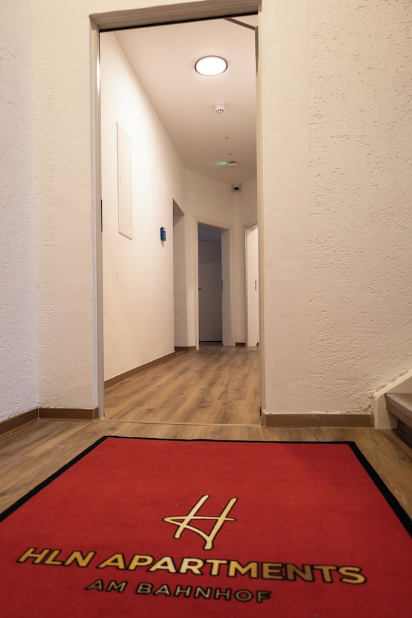 A hallway with a red carpet leading to a door, white walls, and a wooden floor. - HLN APARTMENTS (Saarbrücken)